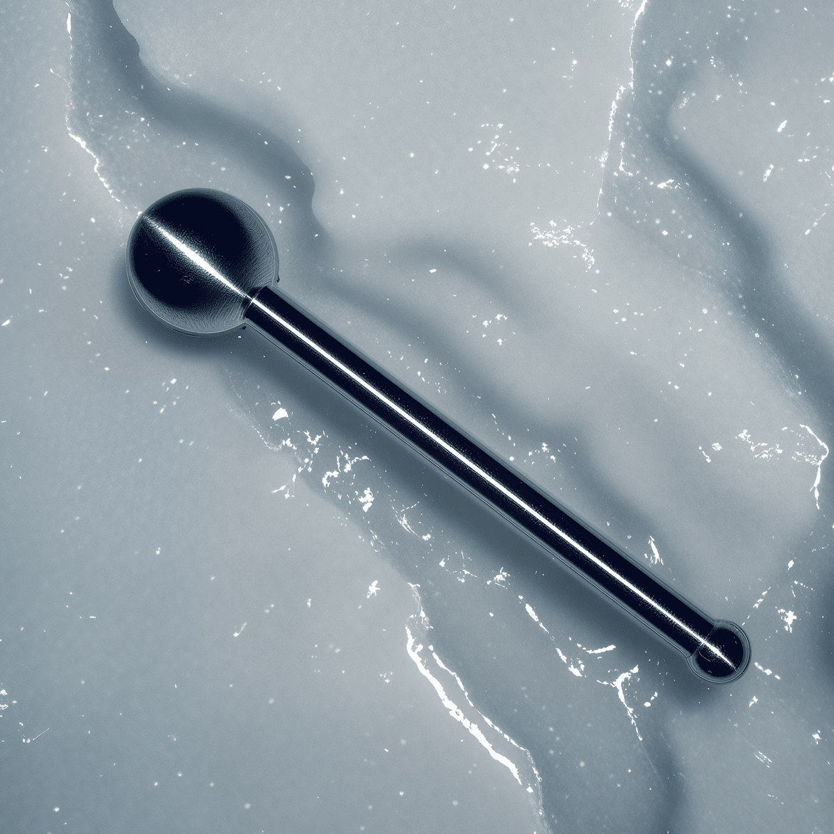 A polished metallic barbell-style jewelry piece with a spherical end and a slim cylindrical shaft, photographed diagonally on a textured light gray surface.