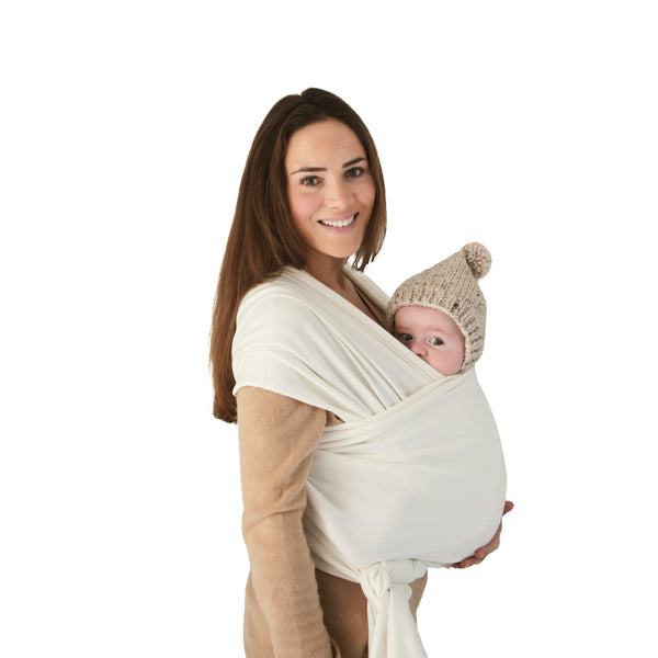 A woman smiles holding a baby in the mushie Baby Wrap. The baby, in a knitted hat, rests snug against her chest. Both face forward against a white background, highlighting the breathable comfort of mushie's Baby Wrap.