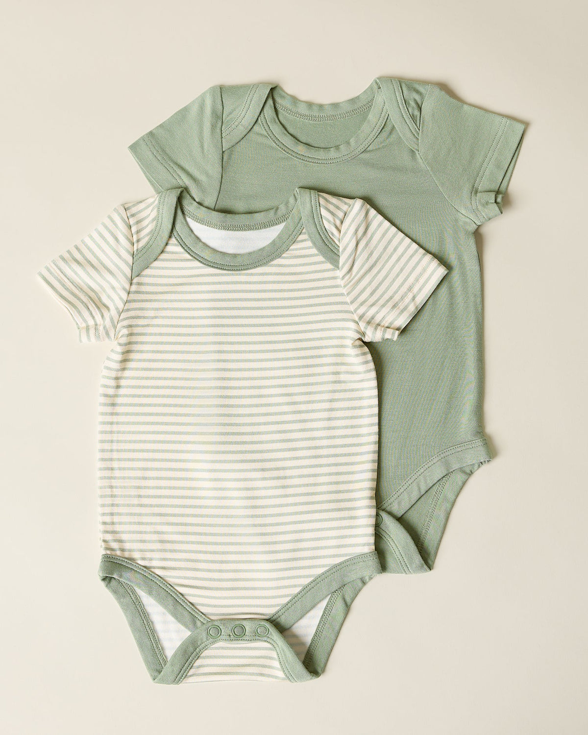 Two green baby short sleeve bodysuits with sage green, one plain and one striped, on a beige background.